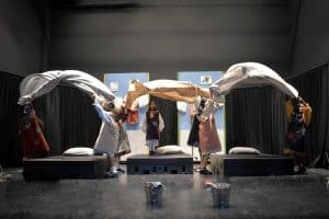 Young actors on stage performing a scene from ‘Annie’ where they are energetically waving bed sheets in the air. The stage is set to resemble an orphanage with props such as beds and buckets, creating a lively and engaging performance atmosphere.