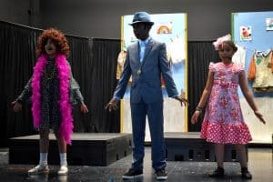 Three young actors on stage performing ‘Easy Street’ from ‘Annie,’ dressed in costumes that include a feather boa, a suit, and a polka-dot dress. The backdrop shows laundry hanging on a line, reflecting the era and setting of the musical.