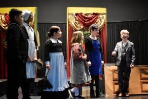 A group of young actors dressed as various characters from ‘Annie’ stand together on stage. The scene appears to be set in a mansion, with actors dressed as servants and other characters, creating a sense of the ensemble cast working together.