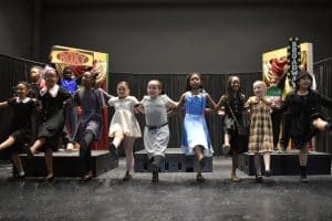 The young cast of ‘Annie’ performs a high-energy dance number, standing in a line with arms around each other, kicking their legs in unison. The stage is decorated with signs for ‘Roxy’ and ‘Broadway,’ adding to the New York City theme of the musical.
